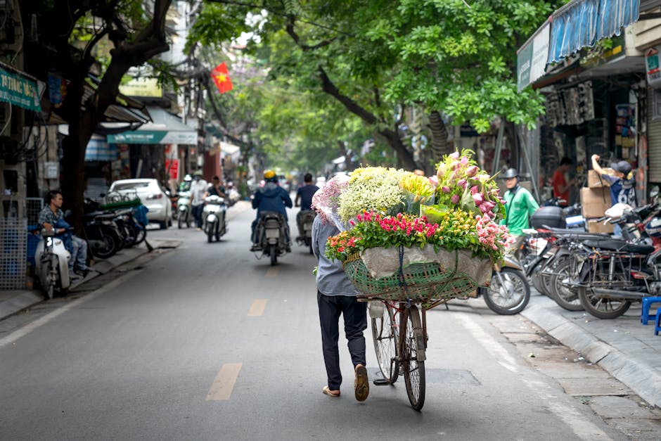 A street scene featuring a street vendor pushing a bicycle cart loaded with fresh, colorful flowers including pink roses, white hydrangeas, and yellow lilies, wrapped in decorative paper and tied with