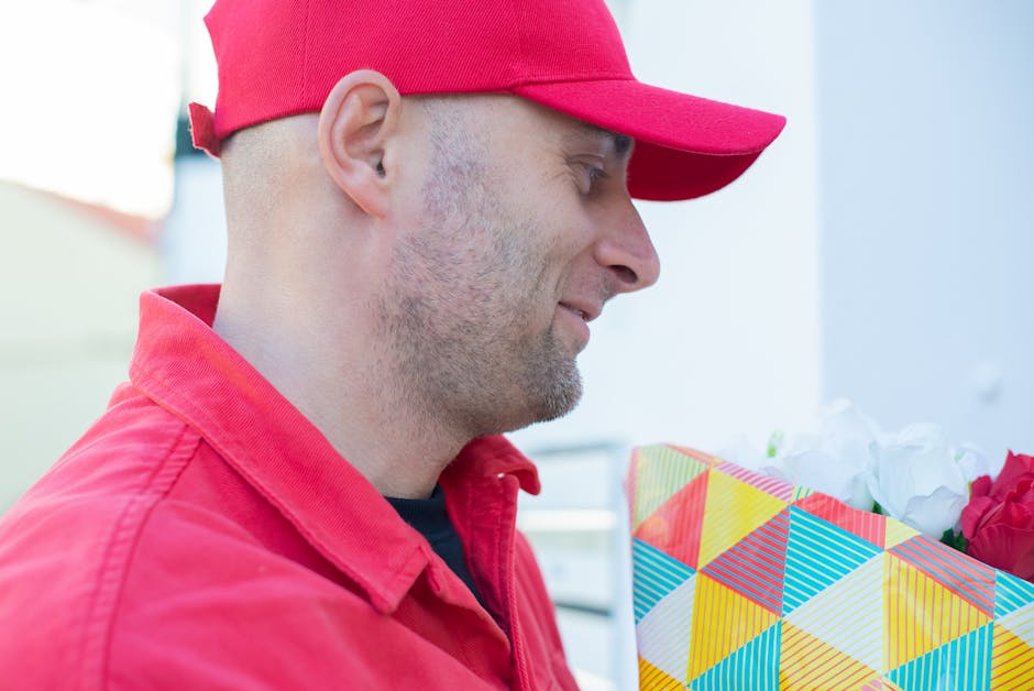 A man wearing a red cap and red jacket is holding a colorful gift box with a triangular pattern in shades of yellow, orange, blue, and green, wrapped with white tissue paper and containing red and whi