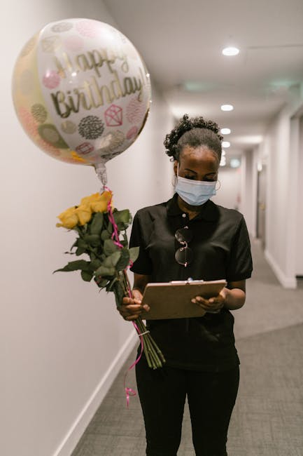 A woman with dark, curly hair wearing a light blue face mask and sunglasses hanging from her collar stands in a well-lit corridor, holding a bouquet of yellow roses tied with a pink ribbon. She is als