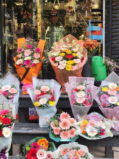 A display of various fresh floral bouquets arranged outdoors in front of a flower shop window. The bouquets feature a mix of pink, yellow, white, and red flowers, including daisies and chrysanthemums,