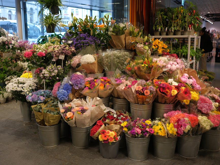 A vibrant display of fresh flower bouquets arranged in metal buckets and wrapped in brown craft paper, featuring a variety of colorful blooms including pink gerbera daisies, yellow sunflowers, purple