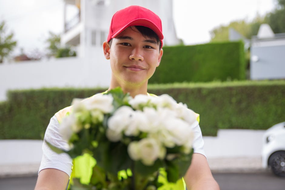 A delivery person wearing a red cap and high-visibility vest holds out a floral gift of white roses and green foliage, arranged in a compact bouquet. The flowers have fresh, soft petals and sturdy gre
