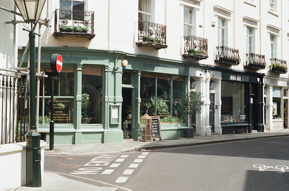 A charming florist shop with a pastel green exterior and large glass windows displaying vibrant flower arrangements inside. The shop has decorative brackets and a small chalkboard sign outside, indica
