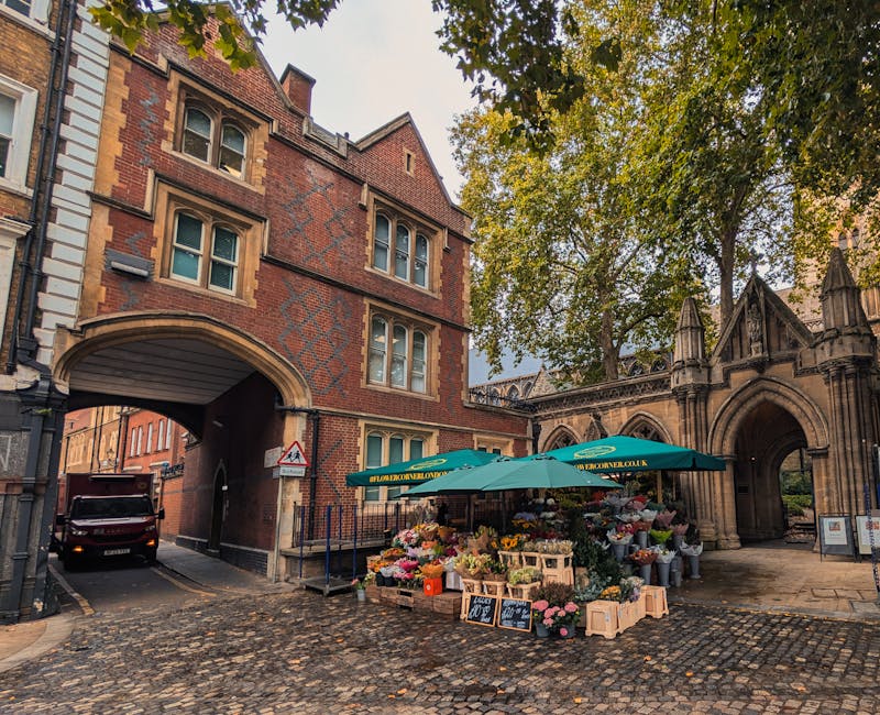 A vibrant flower stall situated on a cobblestone street beneath large green umbrellas, offering a variety of fresh floral arrangements including colorful roses, tulips, peonies, and mixed seasonal blo