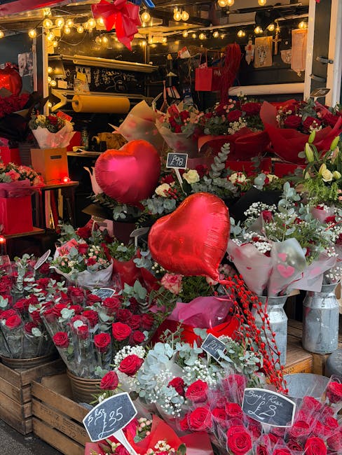 A vibrant floral display at a flower shop showcasing an abundance of red and white roses, several large heart-shaped balloons, and mixed greenery. The arrangement includes wrapped bouquets with pink a
