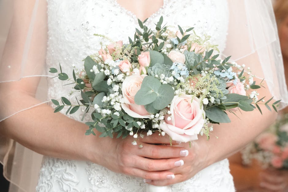 A person wearing a white, textured wedding dress holds a delicate bouquet arrangement consisting of soft pink roses, pale blue and white accent flowers, and green foliage including eucalyptus leaves.